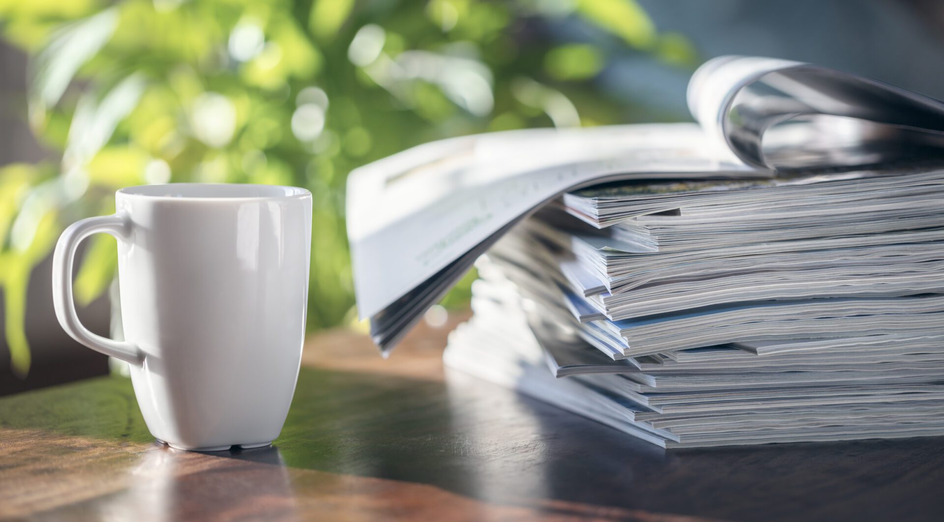 Stack of magazines and white coffee cup on wooden table with blurred