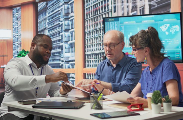 Three colleagues discussing documents around a desk with a digital data screen