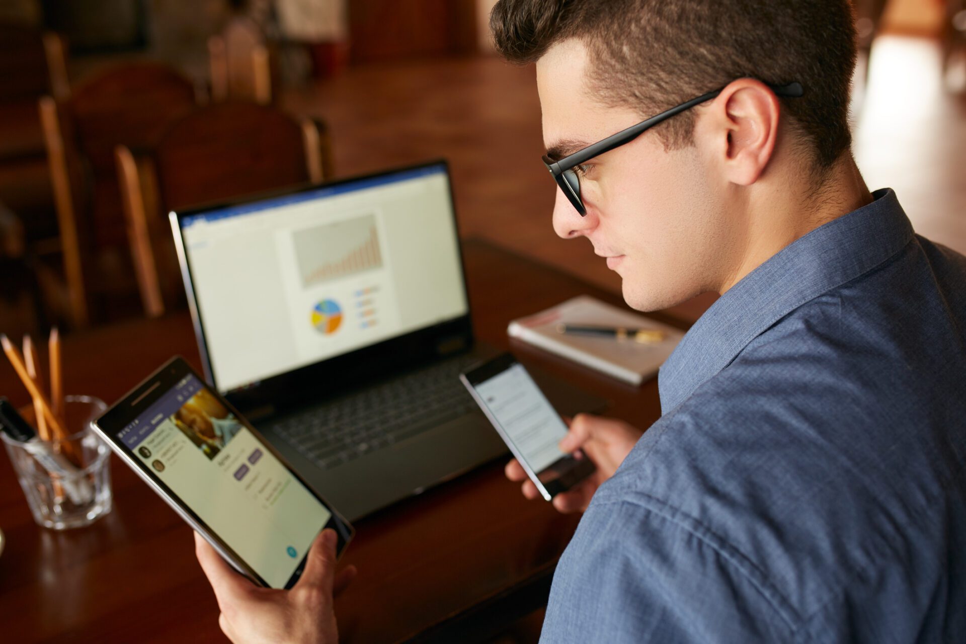 Man in glasses working with multiple electronic internet devices. Freelancer businessman has laptop and smartphone in hands and laptop on table with charts on screen. Multitasking theme