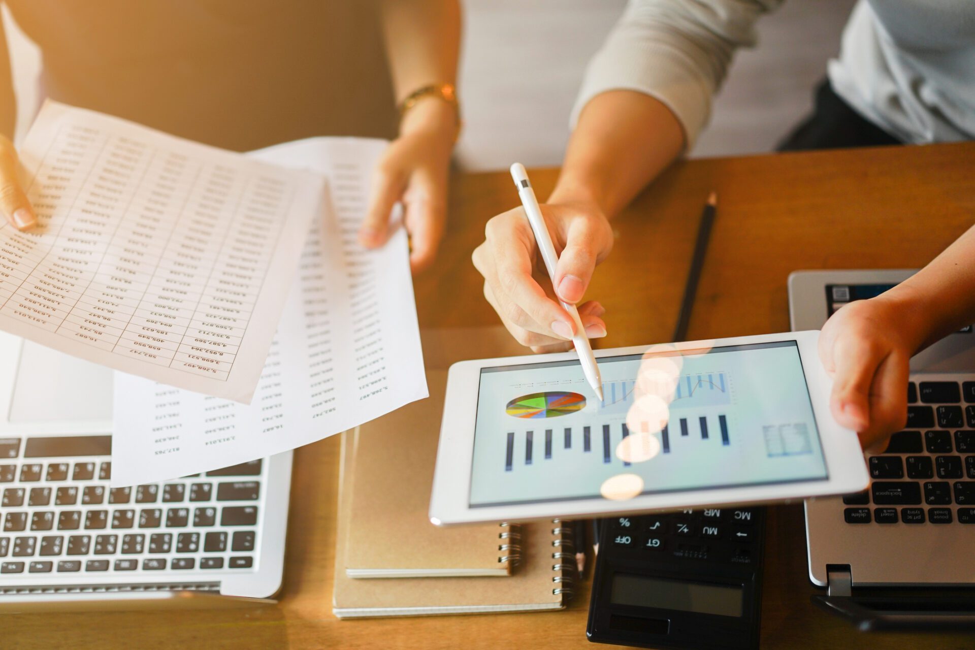 Business professionals analyzing financial data on a tablet and printed sheets, working on laptops at a wooden desk